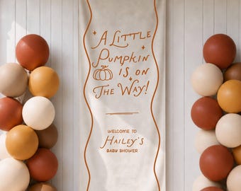 May include: A vertical off-white fabric banner with a wavy orange border and the text A LITTLE PUMPKIN IS ON THE WAY! WELCOME TO HAILEY'S BABY SHOWER in orange. Two balloon columns in shades of cream, tan, and burnt orange flank the banner.