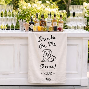 May include: A white linen towel draped over a bar, featuring a black line drawing of a golden retriever and the text: Drinks On Me, Cheers!, XOXO, Olly. The towel is positioned in front of various liquor bottles, glassware, and cocktail garnishes.