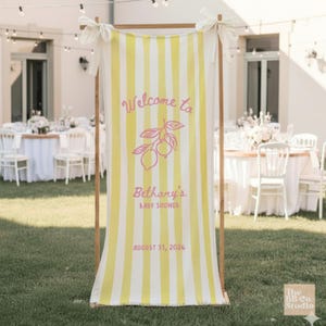 May include: A vertical fabric banner with yellow and white stripes, suspended from a wooden frame with white ribbons. The banner features pink text reading Welcome to Bethany's BABY SHOWER AUGUST 31, 2026, and a pink line drawing of two lemons.