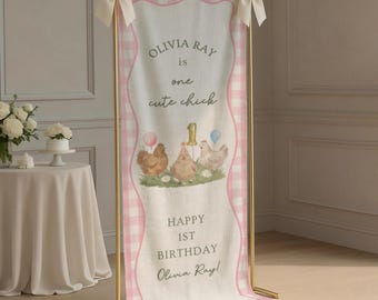 May include: A vertical fabric banner featuring a pink and white gingham border, displayed on a gold metal stand with cream bows. The banner displays the text OLIVIA RAY is one cute chick, a gold number 1, three illustrated chicks, and HAPPY 1ST BIRTHDAY.