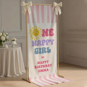 May include: A vertical fabric banner hanging on a gold metal stand with white bows at the top corners. The banner features pink vertical stripes, a yellow sun icon, and the text "ONE HAPPY GIRL" in pink, purple, and blue, plus "HAPPY BIRTHDAY EMMA".