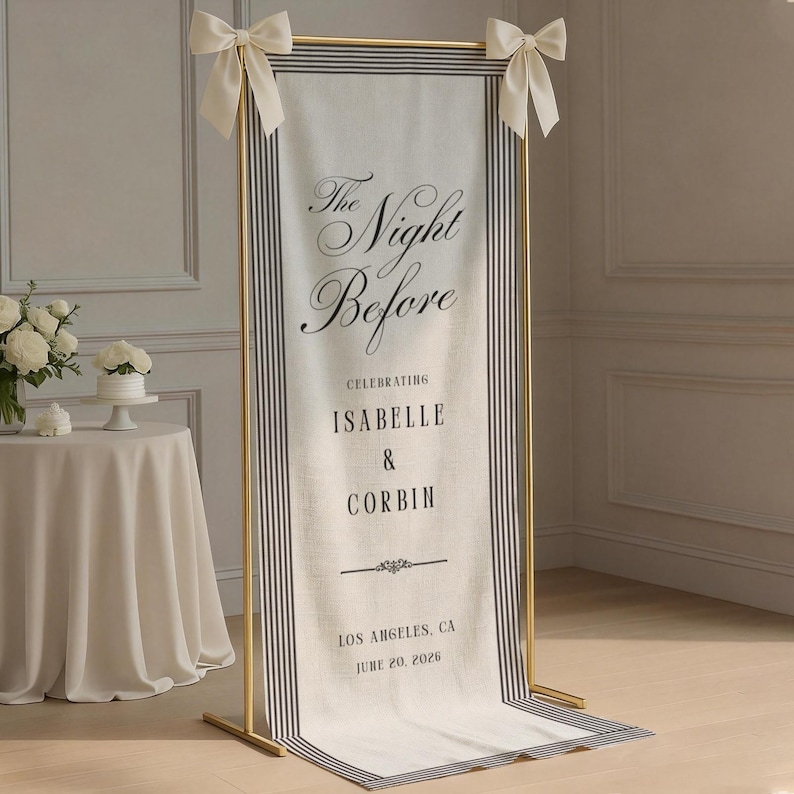May include: A tall, rectangular fabric banner suspended on a gold metal frame with cream-colored bows at the top corners. The banner features black vertical stripes on the sides and black text reading The Night Before, CELEBRATING ISABELLE & CORBIN, LOS ANGELES, CA, JUNE 20, 2026.