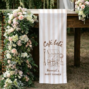 May include: A beige and white striped table runner with the text "Café bébé" and "Hannah's BABY SHOWER" in brown script. The design includes a cafe scene with a street lamp, table, and chairs. The runner is draped over a wooden table.
