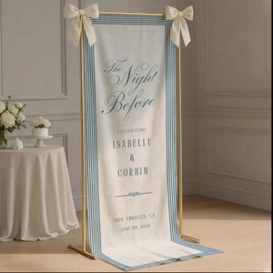 May include: A tall, rectangular fabric banner suspended on a gold metal frame with cream-colored bows at the top corners. The banner features blue vertical stripes along the edges and blue text reading The Night Before, Celebrating Isabelle & Corbin, Los Angeles, CA, June 20, 2026.