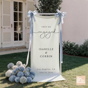 May include: A vertical cream-colored fabric banner suspended on a white frame, featuring the black text "THEY'RE engaged ISABELLE & CORBIN Los Angeles, CA June 20, 2026". The banner is adorned with light blue satin ribbons tied at the top corners.