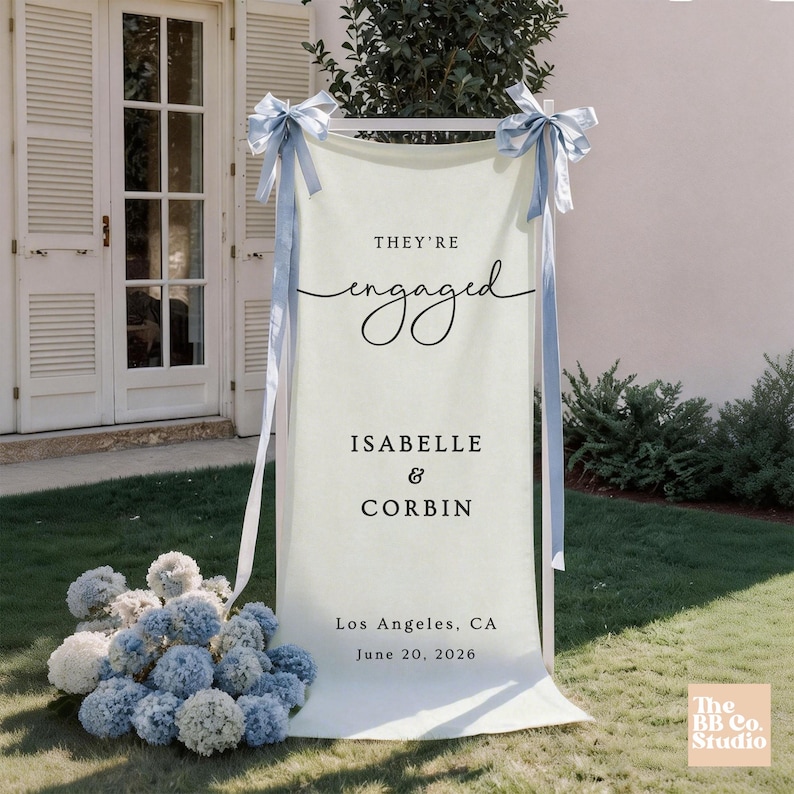 May include: A vertical cream-colored fabric banner suspended on a white frame, featuring the black text "THEY'RE engaged ISABELLE & CORBIN Los Angeles, CA June 20, 2026". The banner is adorned with light blue satin ribbons tied at the top corners.