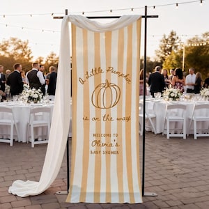 May include: A vertical fabric banner with yellow and white vertical stripes, featuring a pumpkin illustration and the text A Little Pumpkin is on the way WELCOME TO Olivia's BABY SHOWER, draped over a black metal frame stand.