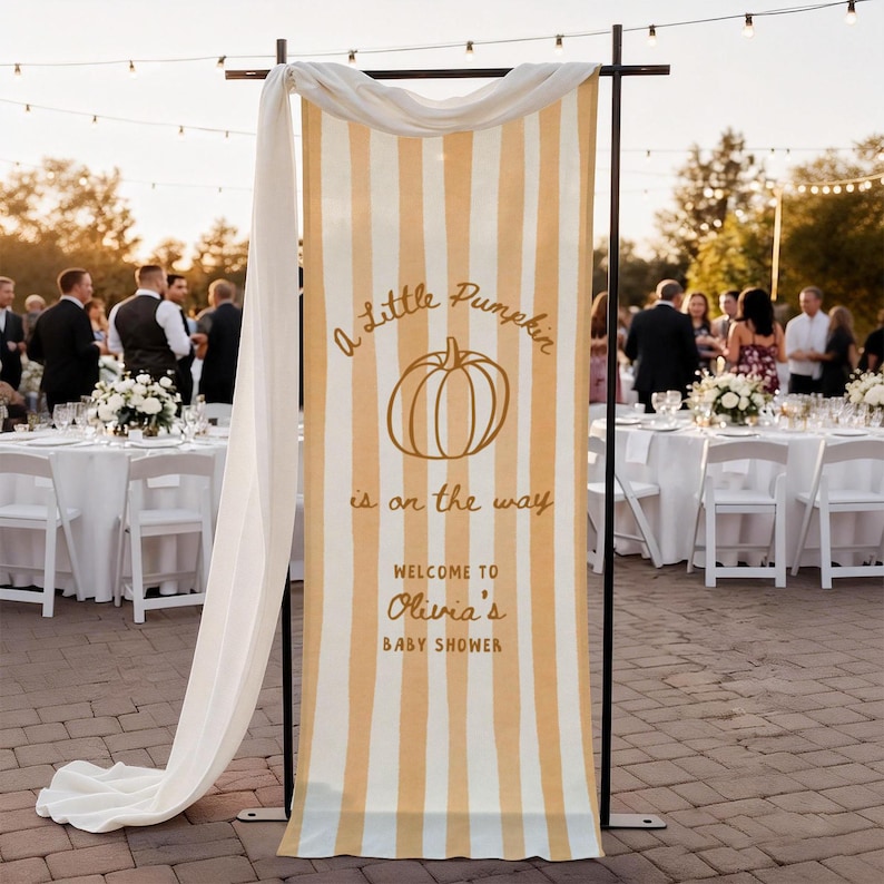 May include: A vertical fabric banner with yellow and white vertical stripes, featuring a pumpkin illustration and the text A Little Pumpkin is on the way WELCOME TO Olivia's BABY SHOWER, draped over a black metal frame stand.