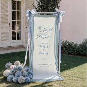 May include: A vertical fabric banner with blue striped borders, tied with light blue ribbons at the top corners. The text reads: The Night Before, CELEBRATING ISABELLE & CORBIN, LOS ANGELES, CA, JUNE 20, 2026. A cluster of blue and white hydrangea flowers sits at the base.