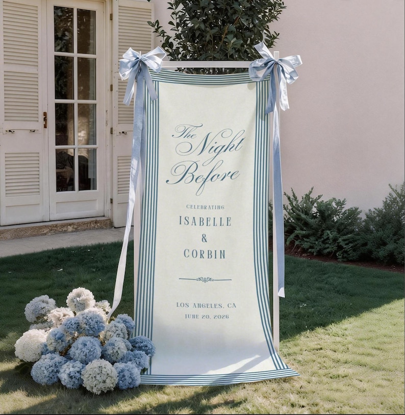 May include: A vertical fabric banner with blue striped borders, tied with light blue ribbons at the top corners. The text reads: The Night Before, CELEBRATING ISABELLE & CORBIN, LOS ANGELES, CA, JUNE 20, 2026. A cluster of blue and white hydrangea flowers sits at the base.