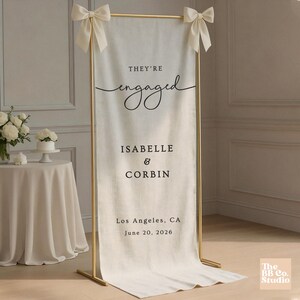 May include: A tall, off-white fabric banner suspended from a gold metal frame with cream-colored bows at the top corners. The banner features black text reading "THEY'RE engaged," "ISABELLE & CORBIN," "Los Angeles, CA," and "June 20, 2026."