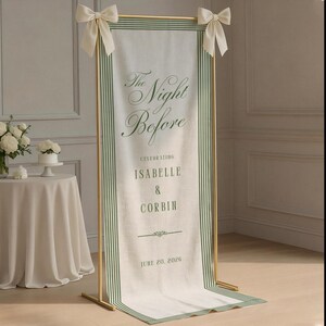 May include: A tall, rectangular fabric banner with green striped borders, suspended from a gold metal frame with cream-colored bows. The banner features the text: The Night Before, CELEBRATING ISABELLE & CORBIN, JUNE 20, 2026, in green lettering.