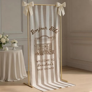May include: A vertical fabric banner with beige and white vertical stripes, suspended on a gold metal frame with cream bows at the top corners. The banner features the brown text Bonjour Bébé, a cafe illustration, and Welcome to Annalisa's Baby Shower.