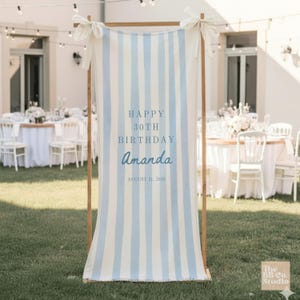 May include: A vertical fabric banner with light blue and white stripes, suspended from a wooden frame with white ribbons. The text reads HAPPY 30TH BIRTHDAY Amanda AUGUST 31, 2026 in blue lettering. The banner is displayed outdoors on a grassy lawn.
