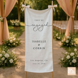 May include: A vertical off-white fabric banner suspended from a wooden frame with tied bows at the top corners. The banner features black printed text reading "THEY'RE engaged", "ISABELLE & CORBIN", "Los Angeles, CA", and "June 20, 2026".