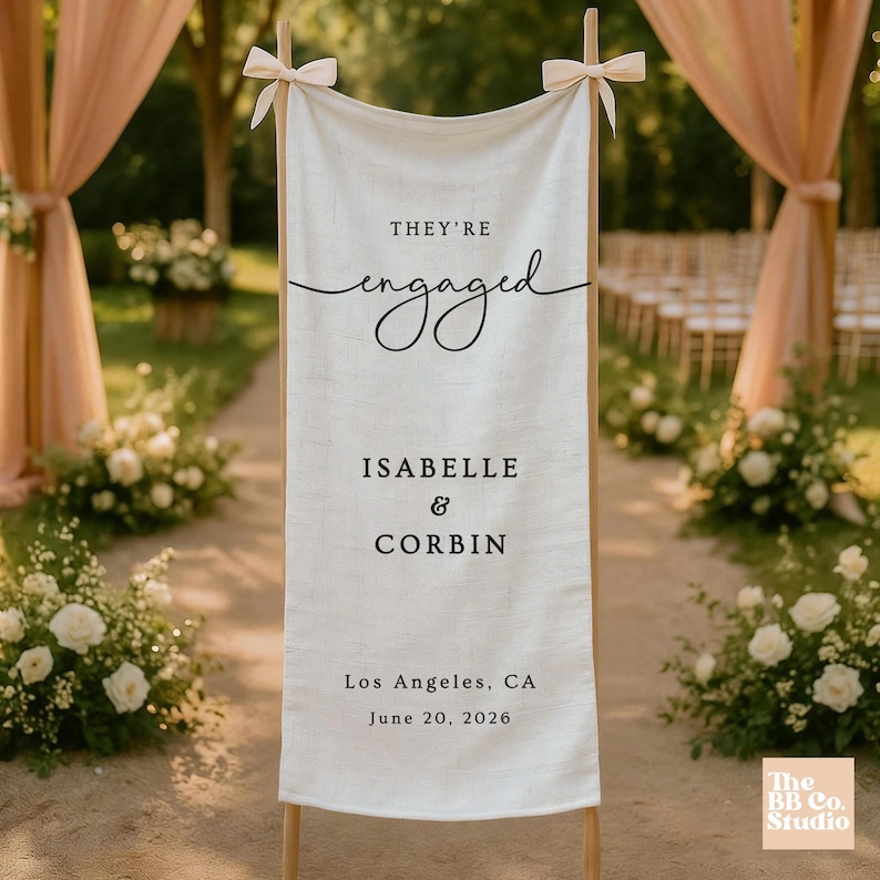 May include: A vertical off-white fabric banner suspended from a wooden frame with tied bows at the top corners. The banner features black printed text reading "THEY'RE engaged", "ISABELLE & CORBIN", "Los Angeles, CA", and "June 20, 2026".