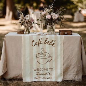May include: A table setting with a beige tablecloth and a striped runner featuring the text "Café bébé" and a coffee cup illustration. The runner says "Welcome to Hannah's Baby Shower". Floral arrangements and decorative items are on the table.