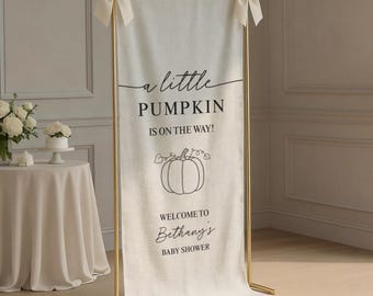 May include: A vertical off-white fabric banner suspended on a gold metal frame with cream-colored bows at the top corners. The banner features black text reading a little PUMPKIN IS ON THE WAY! WELCOME TO Bethany's BABY SHOWER and a pumpkin graphic.