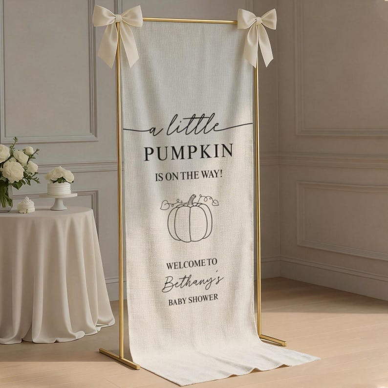 Puede incluir: Una pancarta vertical de tela color hueso suspendida en un marco de metal dorado con lazos color crema en las esquinas superiores. La pancarta presenta texto en negro que dice a little PUMPKIN IS ON THE WAY! WELCOME TO Bethany's BABY SHOWER y un gr&aacute;fico de calabaza.