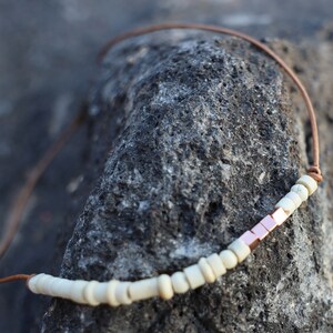May include: A white and copper beaded necklace with a leather cord. The necklace is on a dark gray textured surface.