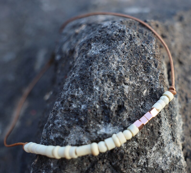 May include: A white and copper beaded necklace with a leather cord. The necklace is on a dark gray textured surface.