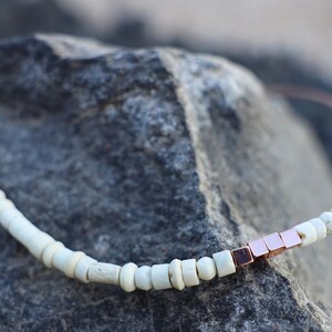 May include: A white and rose gold beaded necklace with a leather cord. The necklace features small white beads and a cluster of rose gold square beads.