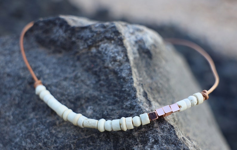 May include: A white and rose gold beaded necklace with a leather cord. The necklace features small white beads and a cluster of rose gold square beads.