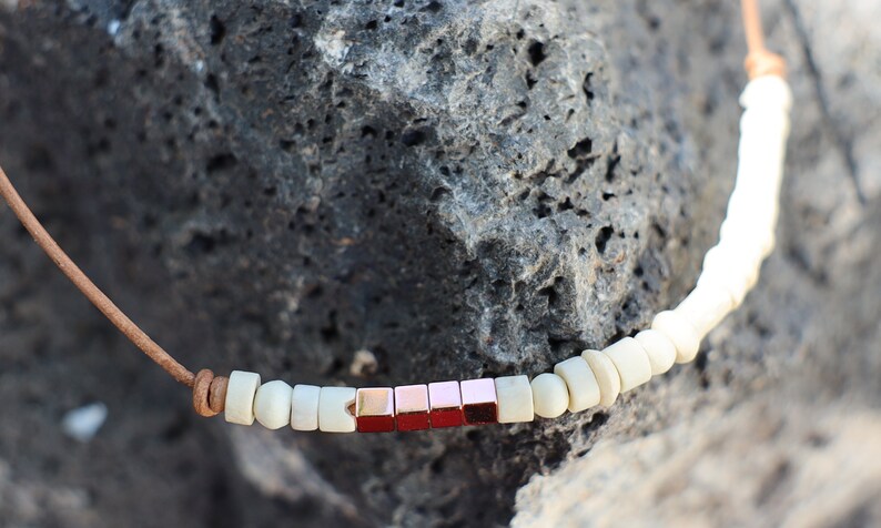 May include: A brown leather cord necklace with white and rose gold square beads.