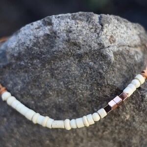 May include: A brown leather cord necklace with white beads and a row of rose gold square beads.