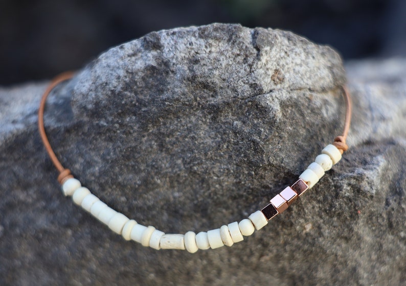 May include: A brown leather cord necklace with white beads and a row of rose gold square beads.