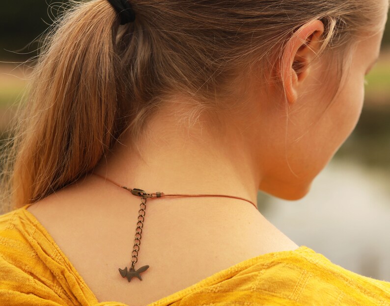 May include: A woman with long blonde hair pulled back in a ponytail is wearing a yellow shirt and a brown leather cord necklace with a small fox charm.