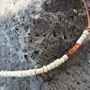 May include: A necklace with white beads and copper accents. The necklace is made of leather cord and is strung with small, round white beads and copper square beads.