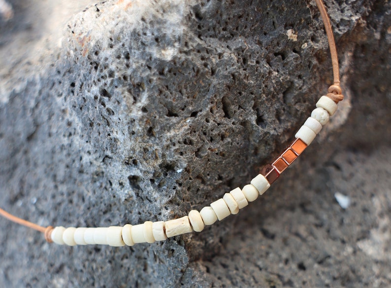 May include: A necklace with white beads and copper accents. The necklace is made of leather cord and is strung with small, round white beads and copper square beads.