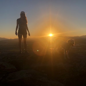 May include: Silhouette of a person and a dog standing on a rocky hilltop, with the sun setting in the background.