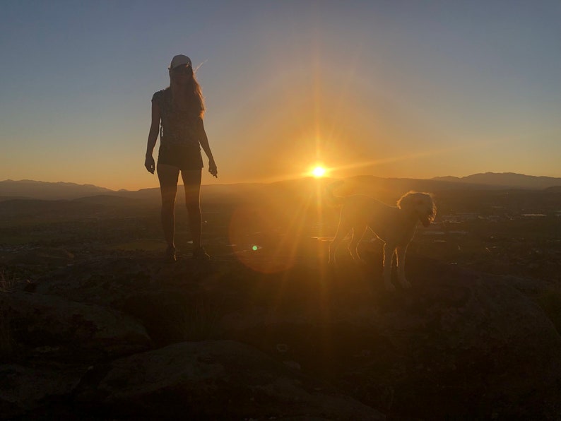 May include: Silhouette of a person and a dog standing on a rocky hilltop, with the sun setting in the background.