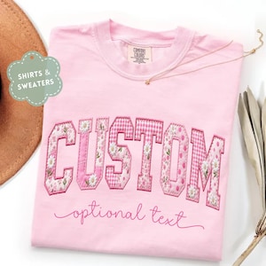 May include: A light pink t-shirt featuring the word CUSTOM in large, block-style letters with varied floral, gingham, and polka dot patterns. Below the block letters, the phrase optional text is written in a cursive, dark pink script font.