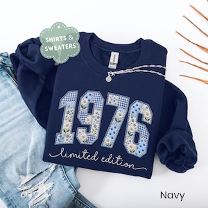 May include: A navy blue crewneck sweatshirt featuring the year 1976 in large, appliqued numbers with a light blue floral and gingham patchwork pattern. Below the numbers, the text "limited edition" is embroidered in white script.