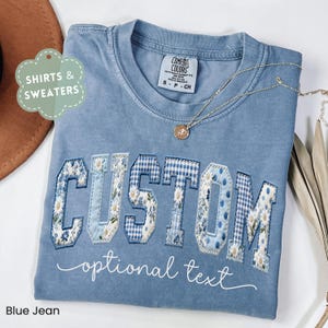 May include: Blue jean colored t-shirt with the word "CUSTOM" in a collegiate style, featuring floral, gingham, and polka dot patterns. The phrase "optional text" is below the word "CUSTOM". A gold necklace with a pendant is on the shirt.