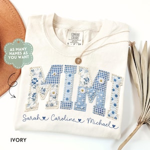 May include: Ivory-colored t-shirt with the word "MIMI" in blue patchwork letters. The letters feature floral, gingham, and polka dot patterns. Below the name are three names: Sarah, Caroline, and Michael. A gold necklace and a brown hat are also visible.