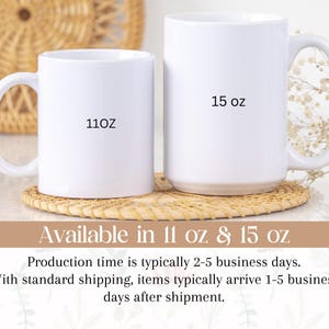 May include: Two white ceramic mugs, one marked "11oz" and the other "15 oz", are displayed on a woven coaster. The mugs have large handles and a glossy finish. The background features a woven basket and floral decorations.