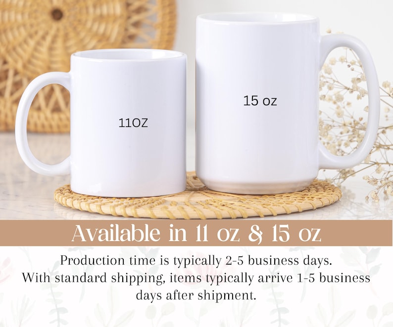 May include: Two white ceramic mugs, one marked "11oz" and the other "15 oz", are displayed on a woven coaster. The mugs have large handles and a glossy finish. The background features a woven basket and floral decorations.