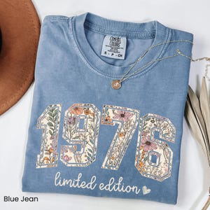 Peut inclure: T-shirt bleu jean avec l'ann&eacute;e "1976" dans un motif floral. Les mots "limited edition" sont imprim&eacute;s en dessous des chiffres. Un collier en or avec un pendentif est drap&eacute; sur le t-shirt.