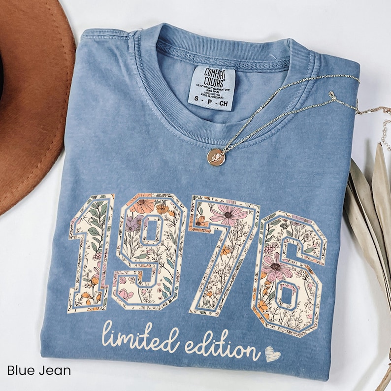 Peut inclure: T-shirt bleu jean avec l'ann&eacute;e "1976" dans un motif floral. Les mots "limited edition" sont imprim&eacute;s en dessous des chiffres. Un collier en or avec un pendentif est drap&eacute; sur le t-shirt.