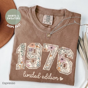 May include: Espresso-colored t-shirt with the year "1976" in a floral design. The words "limited edition" are below the numbers. A silver necklace with a pendant is draped across the shirt.