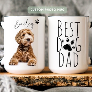 May include: Two white ceramic mugs with black handles. One mug features a photo of a brown curly-haired dog with the name Bailey and a paw print. The other mug displays the black text BEST DOG DAD with a paw print graphic between the words.
