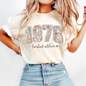 Peut inclure: T-shirt cr&egrave;me avec l'ann&eacute;e "1976" dans un motif floral. Le texte "limited edition" est en dessous. Le t-shirt est port&eacute; avec un jean en denim bleu.