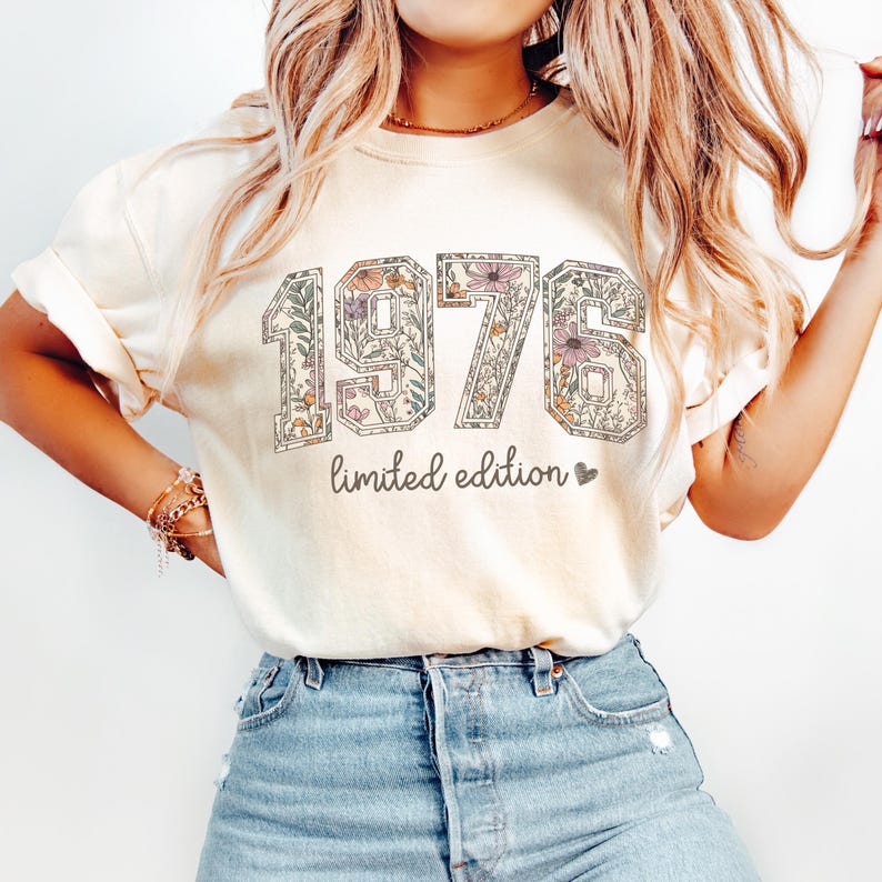 Peut inclure: T-shirt cr&egrave;me avec l'ann&eacute;e "1976" dans un motif floral. Le texte "limited edition" est en dessous. Le t-shirt est port&eacute; avec un jean en denim bleu.