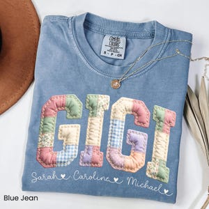 May include: Blue jean Comfort Colors t-shirt with the name "GIGI" in a patchwork design. The shirt also features the names "Sarah", "Caroline", and "Michael" below the name. A gold necklace is draped across the shirt.