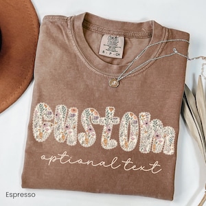 May include: Espresso-colored t-shirt with the word "CUSTOM" in floral letters. The phrase "optional text" is below the word. A silver necklace with a pendant is on the shirt. The shirt is made of Comfort Colors material.