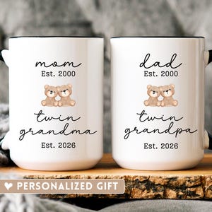 May include: Two white ceramic mugs with black handles and rims. One mug reads "mom, Est. 2000, twin grandma, Est. 2026" and the other reads "dad, Est. 2000, twin grandpa, Est. 2026". Both mugs feature a graphic of two teddy bears. The text "PERSONALIZED GIFT" is at the bottom.